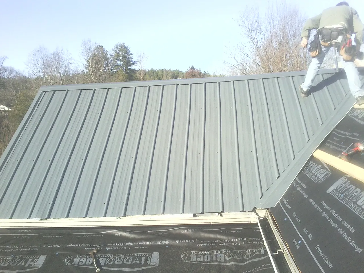 Crew installing dark green standing seam panels over synthetic underlayment in Bolton